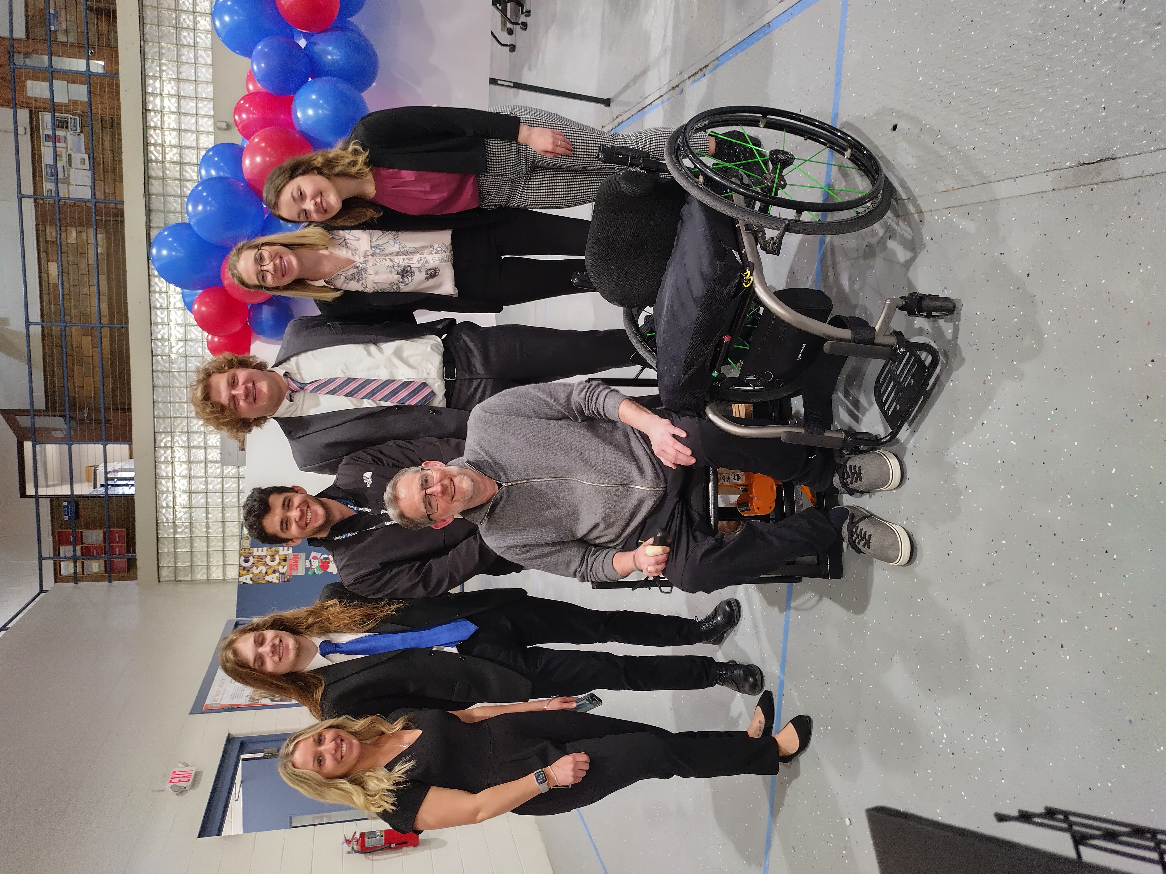 Seven people pose for a photo indoors.  One person sits in a wheelchair next to a prototype wheelchair created by a group of students.