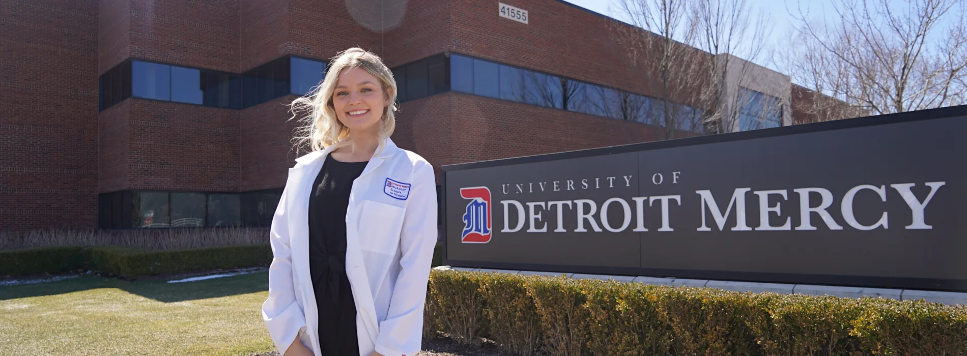 Health Professions student in white coat standing outside Novi location by University sign on sunny day.