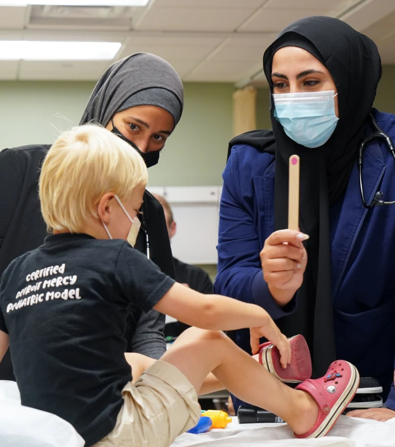 Two PA students work with a child patient. The child wears a shirt that says, Certified Detroit Mercy Pediatric Model