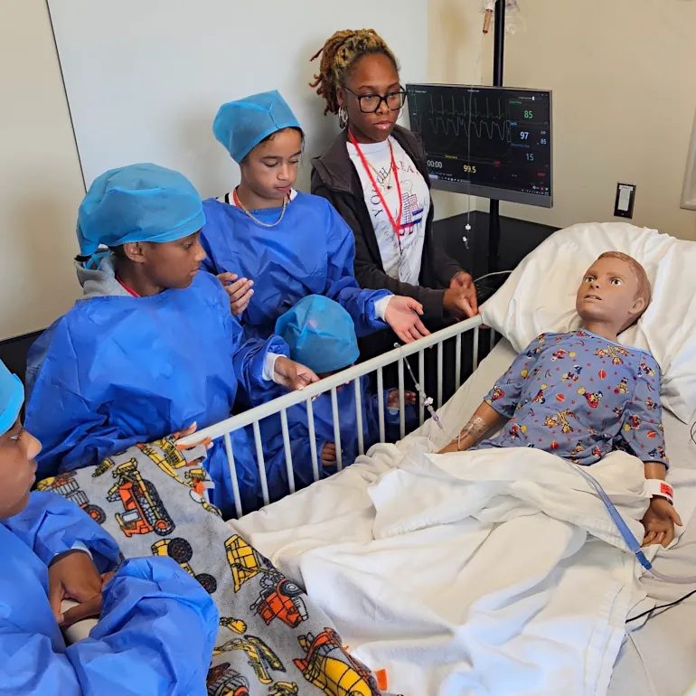 Middle school age campers in hospital garb around a pediatric mannikin in a health professions simulation lab.